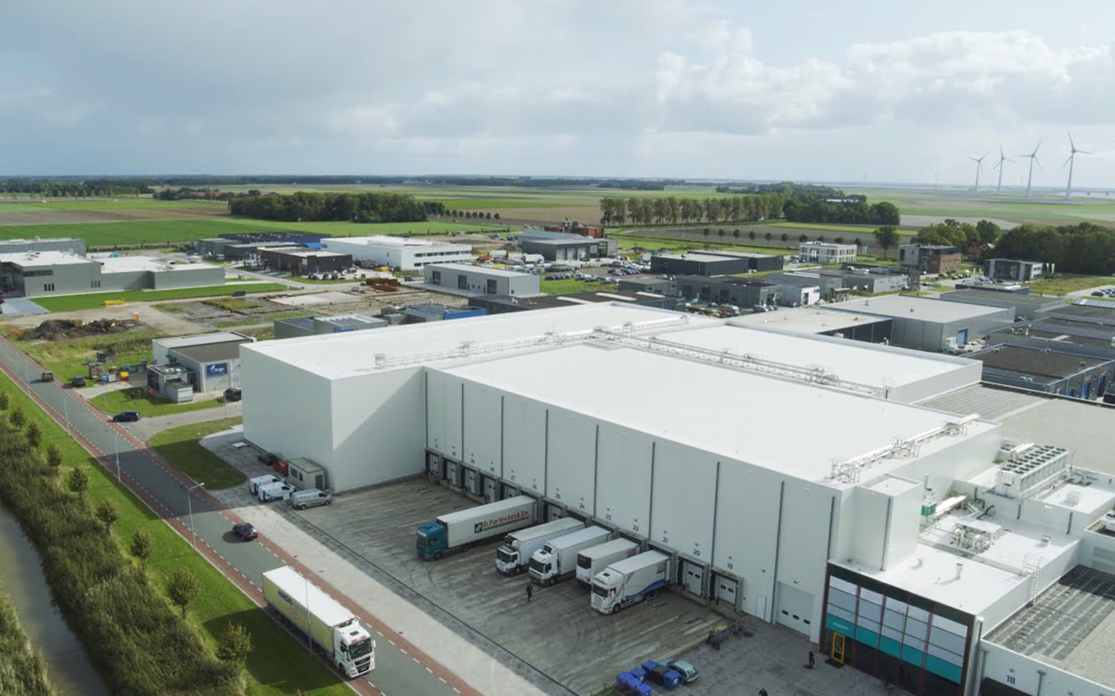 High-bay cold store from the air, trailers at the dock and open polder beyond